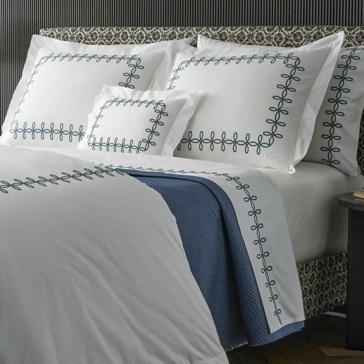 A neatly made bed with Matouk Gordian Knot Bedding, Baltic color embroidery, and a steel blue Alba blanket