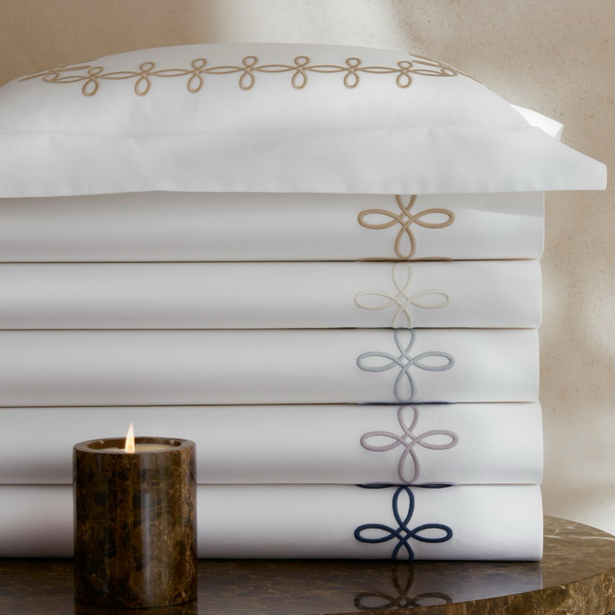 A stack of Matouk Gordian Knot Bedding with a matching pillow, and a lit brown candle in front