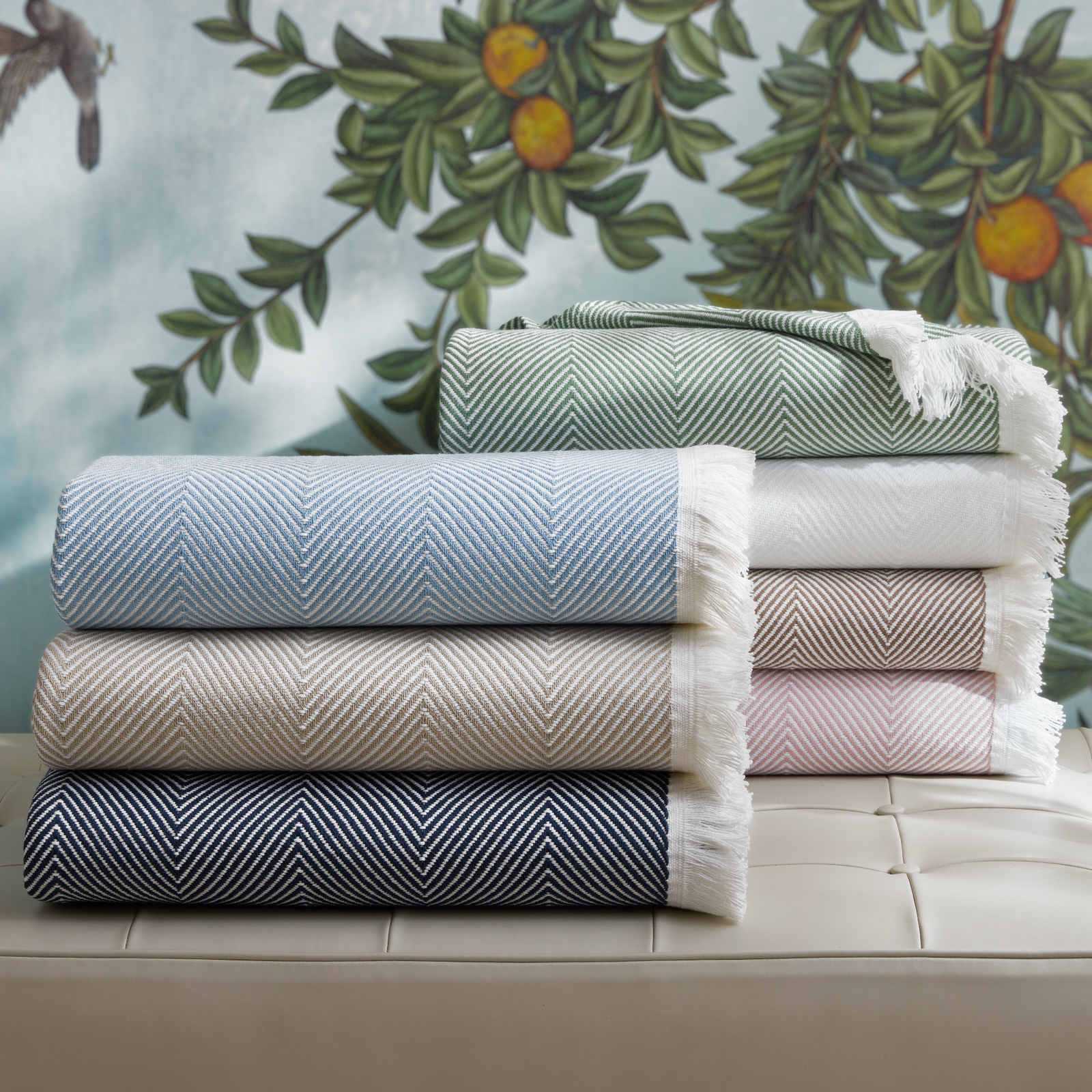 Six folded Matouk Leo herringbone throws in various colors are stacked on a beige cushioned bench.