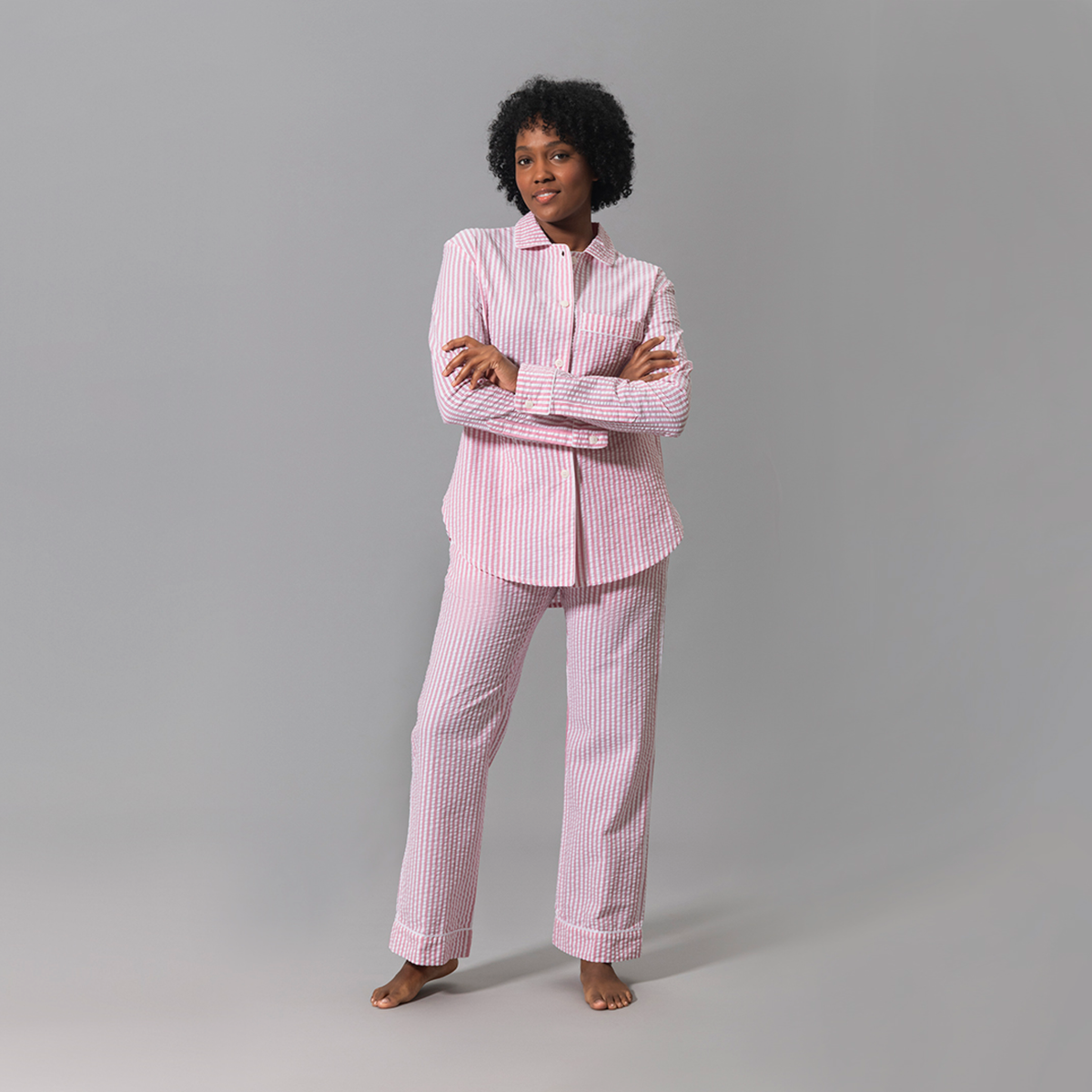 A woman stands barefoot in pink Matouk Matteo Pajama set against a plain gray background