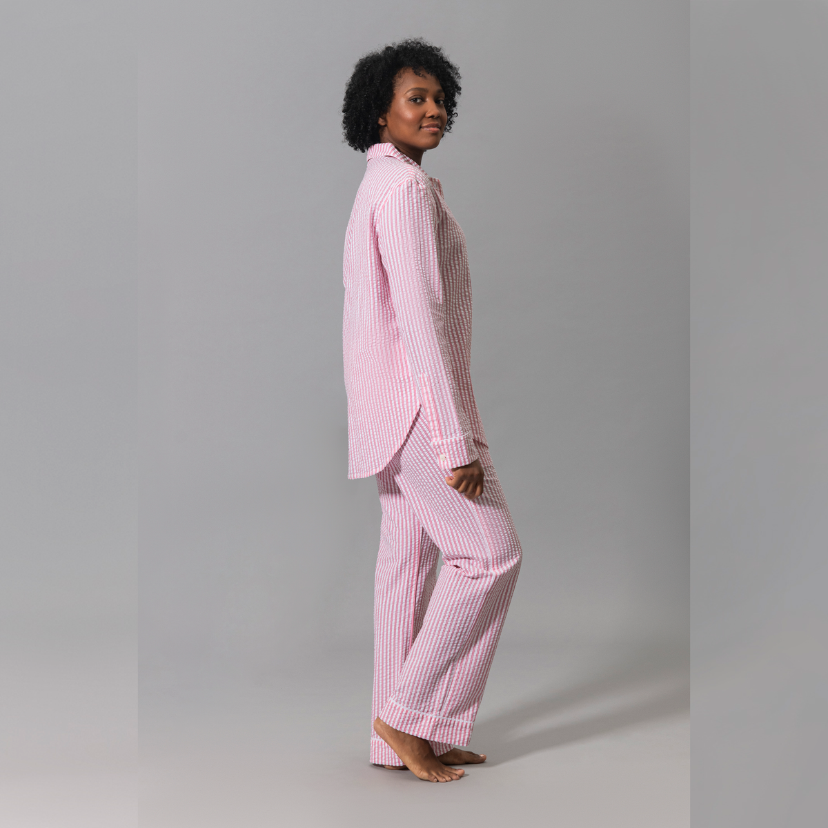 A person in Pink Matouk Matteo Pajama Set stands barefoot on a plain gray background, facing slightly to the side