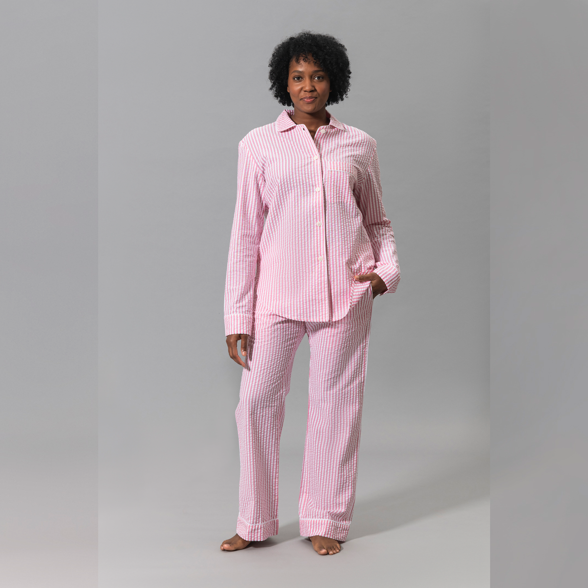 A woman stands barefoot in pink Matouk Matteo Pajama Set against a plain gray background
