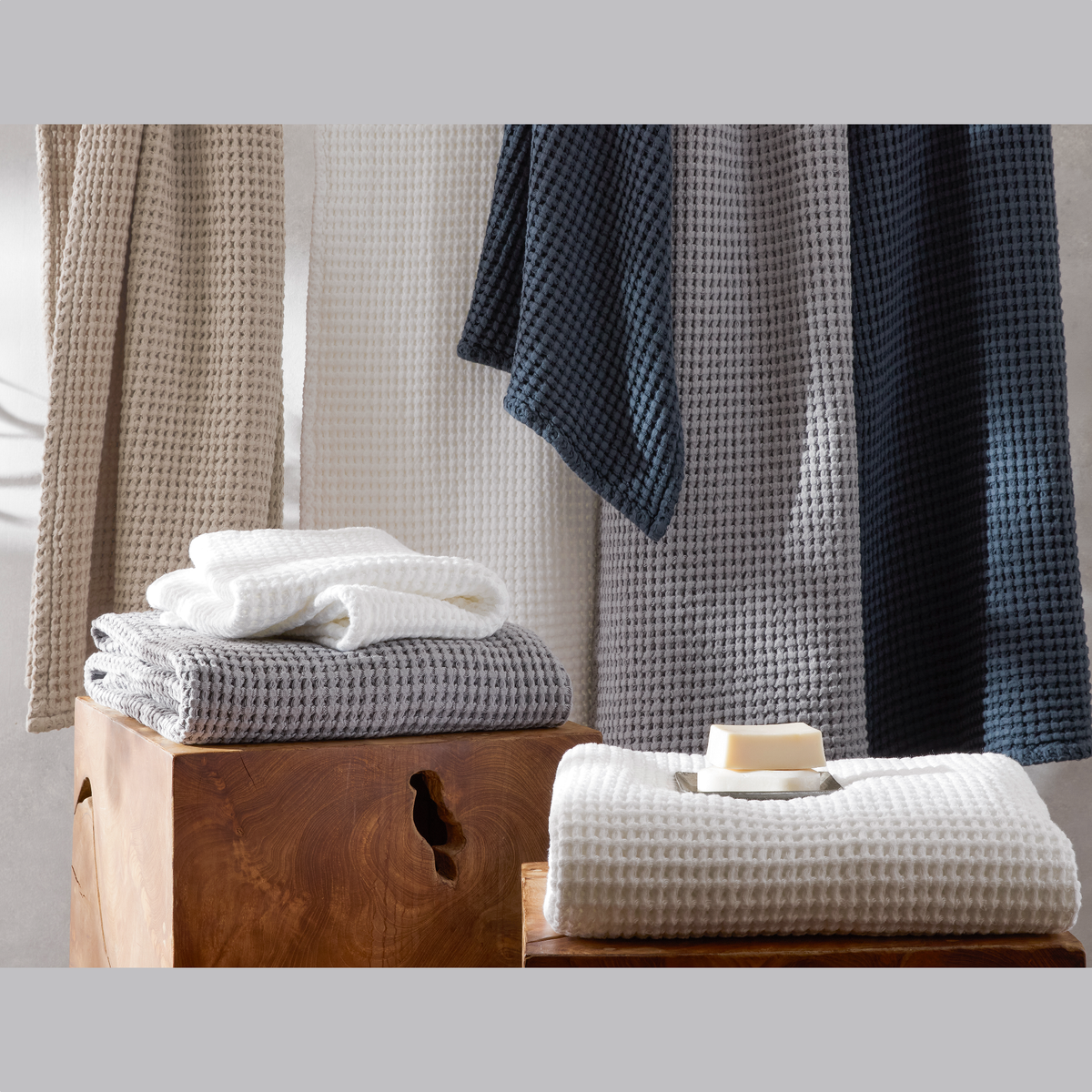 Five Matouk waffle towels in neutral hues hang and fold by a stool, showcasing their signature honeycomb weave.