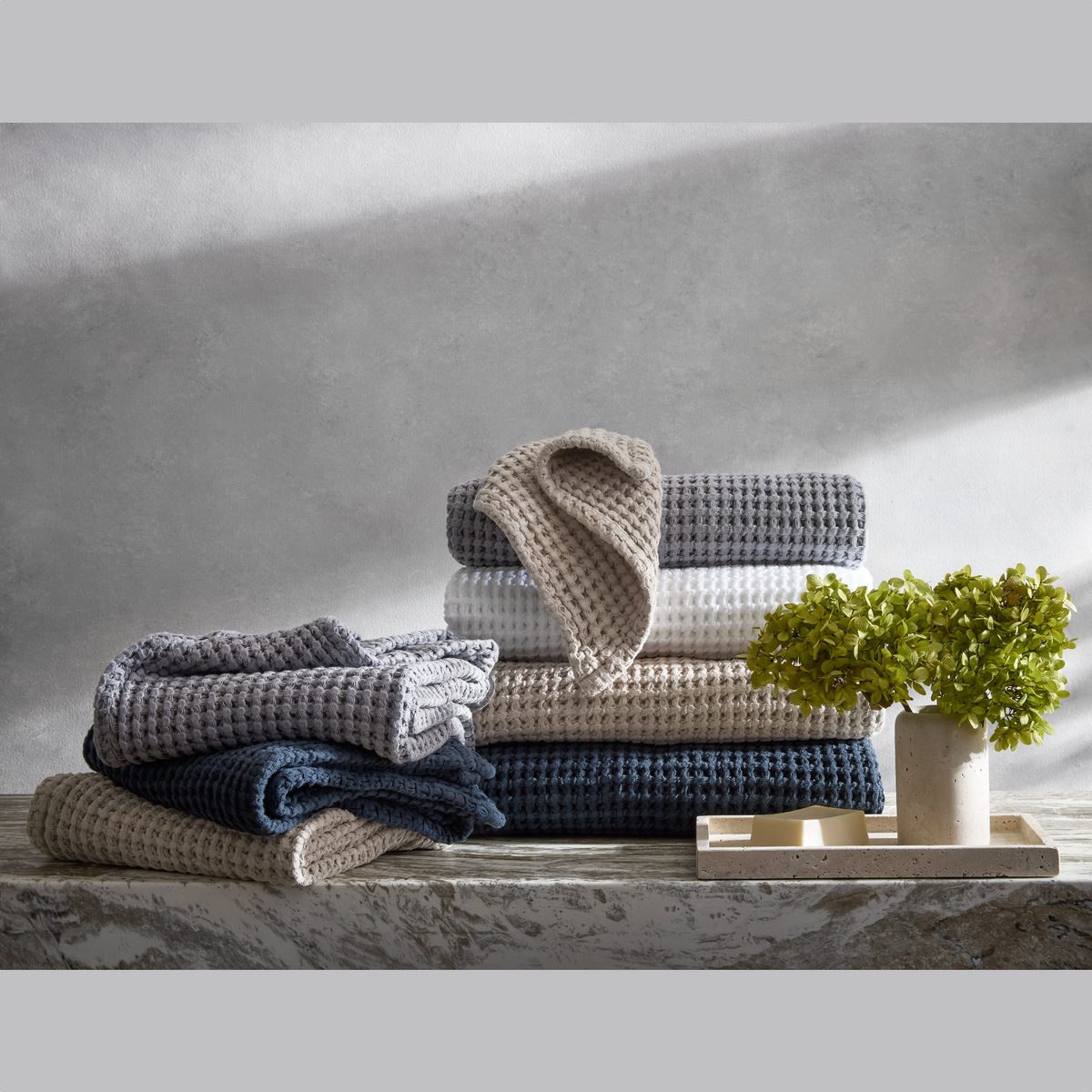 Stacked Matouk waffle towels in honeycomb weave, next to a tray with a potted plant and soap on a marble surface.
