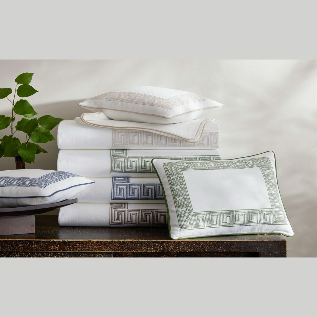 A stack of luxury cotton coverlet and Greek key bedding by Matouk Schumacher Ophelia Pique on a dark wooden surface.