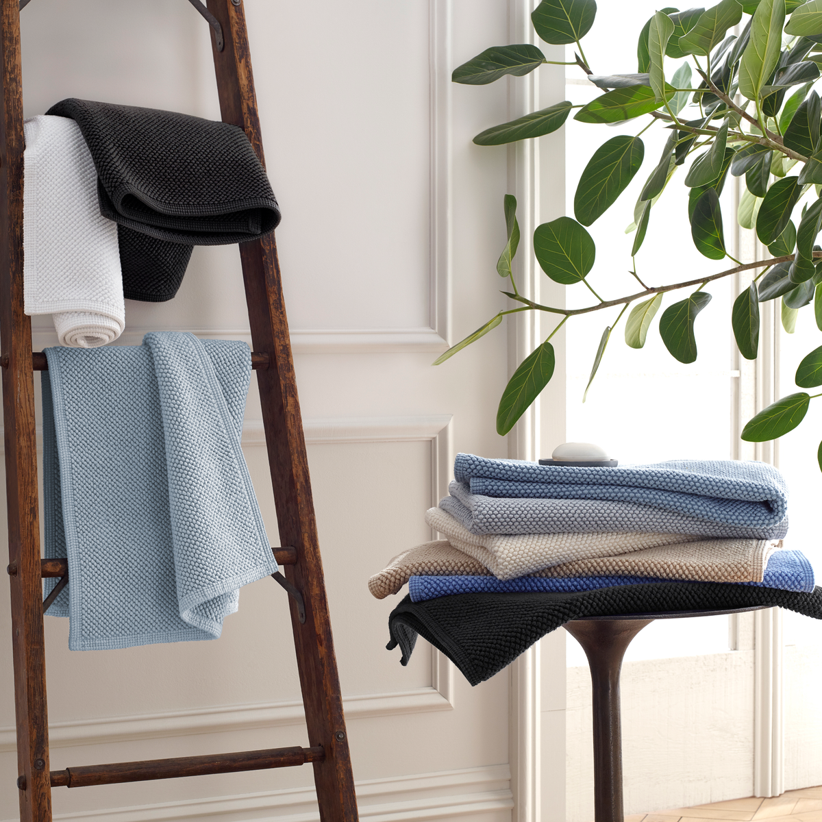 Folded Matouk Reverie bath rugs are displayed on a wooden ladder and small round table indoors