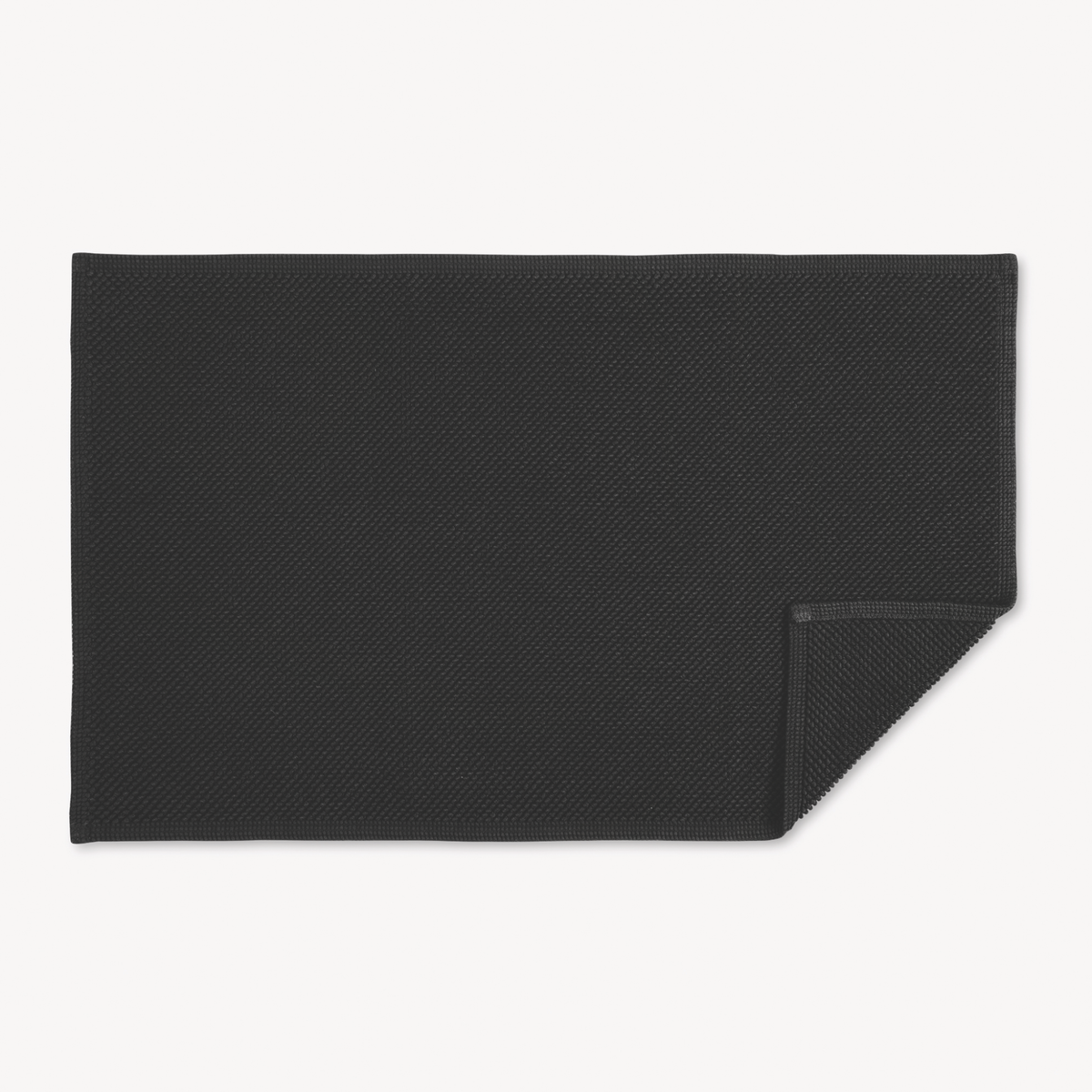 A black textured rectangular Matouk Reverie bath rug with non-slip backing, one corner slightly folded over on white background