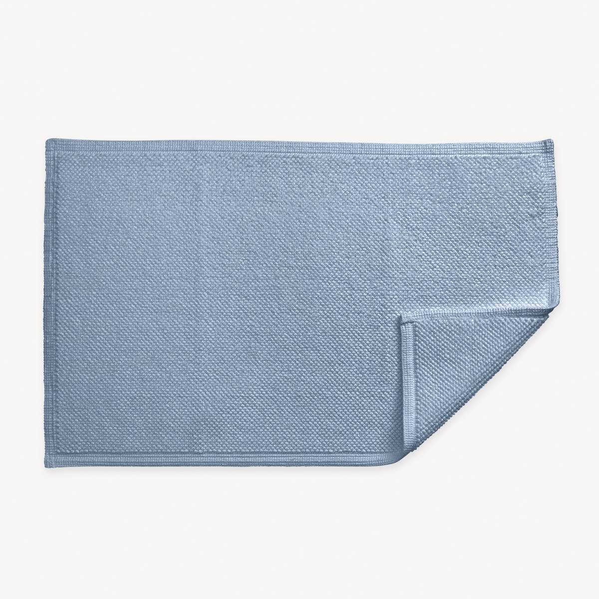 A hazy blue textured rectangular Matouk Reverie bath rug with non-slip backing, one corner slightly folded over on white background