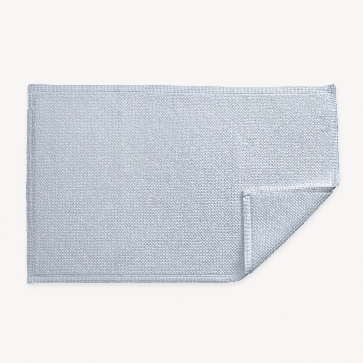 A light blue textured rectangular Matouk Reverie bath rug with non-slip backing, one corner slightly folded over on white background