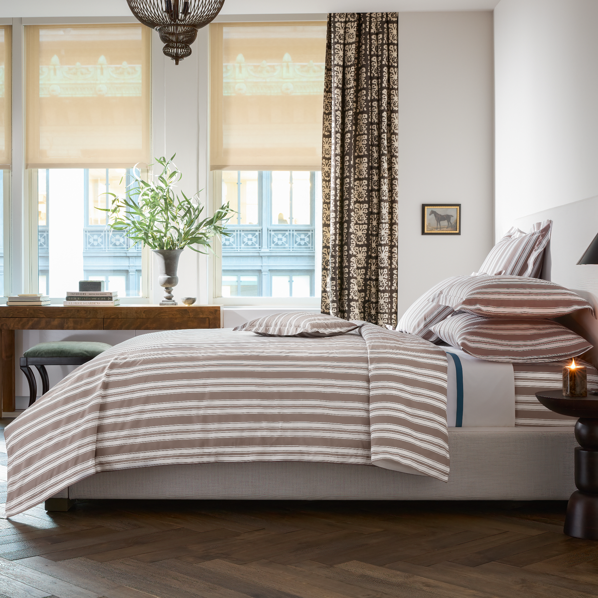 A modern bedroom with Matouk Schumacher Elio Bedding on wooden floors, and a lit candle on the bedside table.
