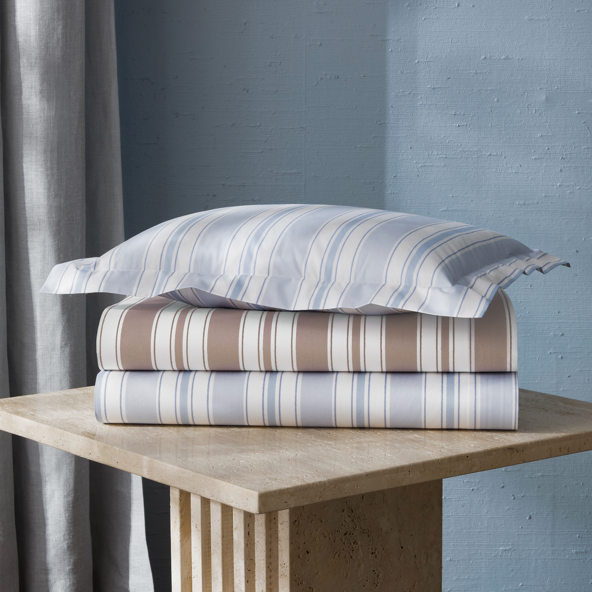 Colors of Matouk Schumacher Elio Bedding on a beige stone pedestal against a blue wall.