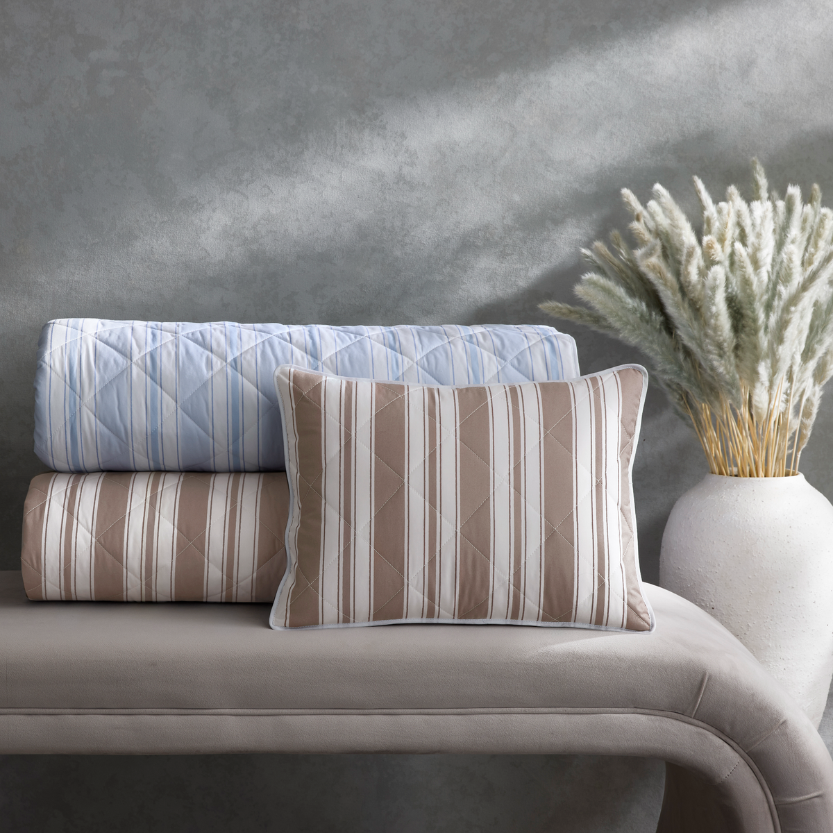 A quilted pillow and two rolled quilts from the Matouk Schumacher Elio Bedding resting in Blue and Mocha