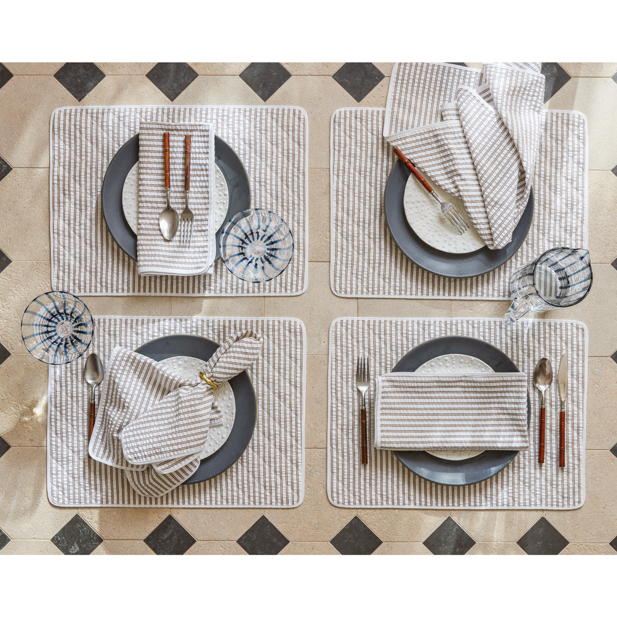 Four place settings with Matouk Matteo table linens — Bark color placemats and napkins, gray plates, cutlery, and glasses on a tiled table