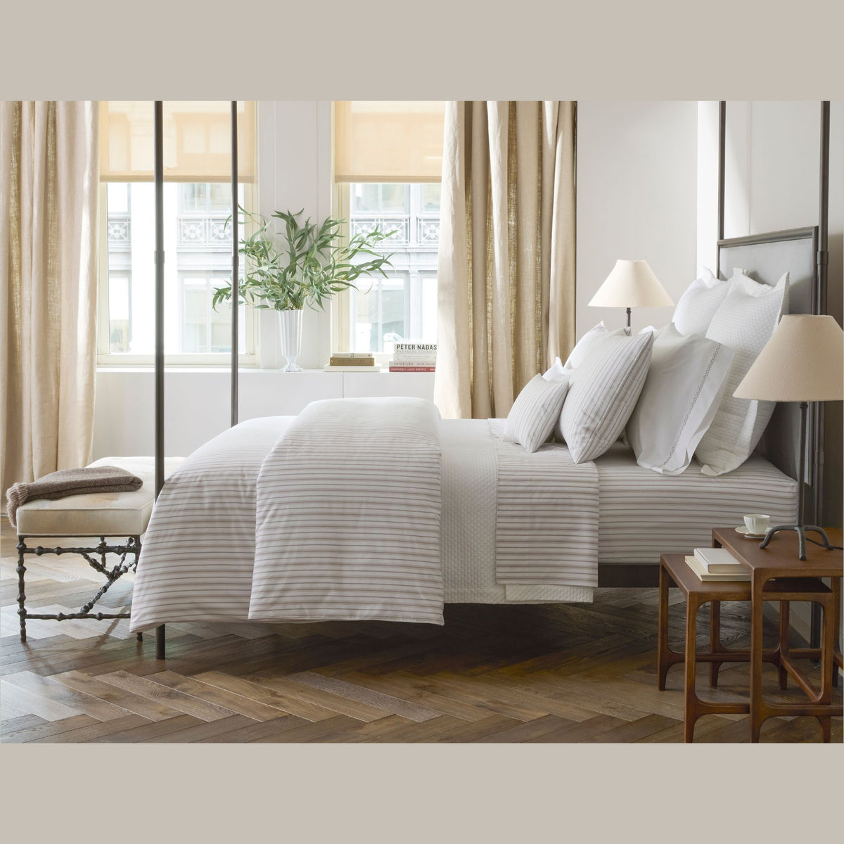 A neatly made bed with Matouk Schumacher Calypso Bedding, two wooden side tables, and a bench in a bright, elegant bedroom.