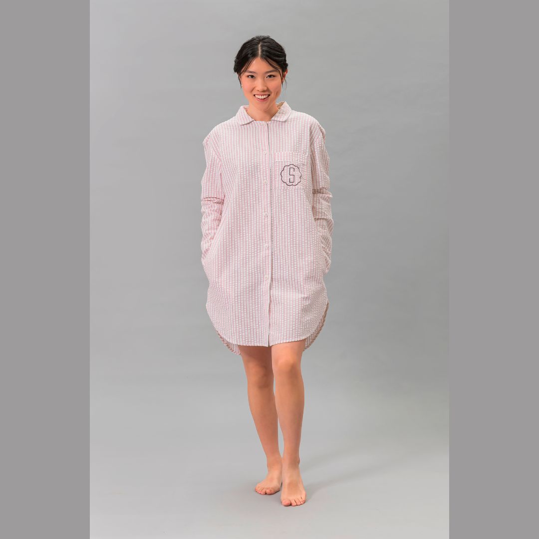 A person stands barefoot on a gray floor, hands in pockets, wearing an oversized sleep shirt in checked luxury cotton.