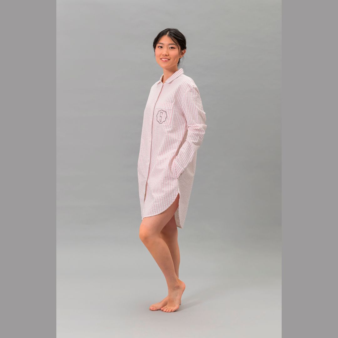 A woman stands barefoot in a Matouk Matteo Sleep Shirt, hands in pockets, against a plain gray background.