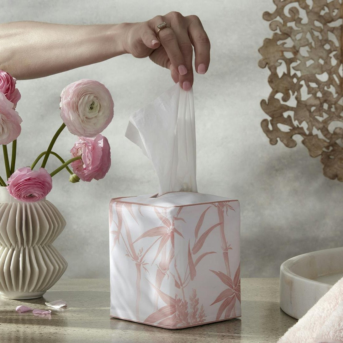 Matouk Dominique Tissue box cover in blush on a table next to a vase of pink flowers.
