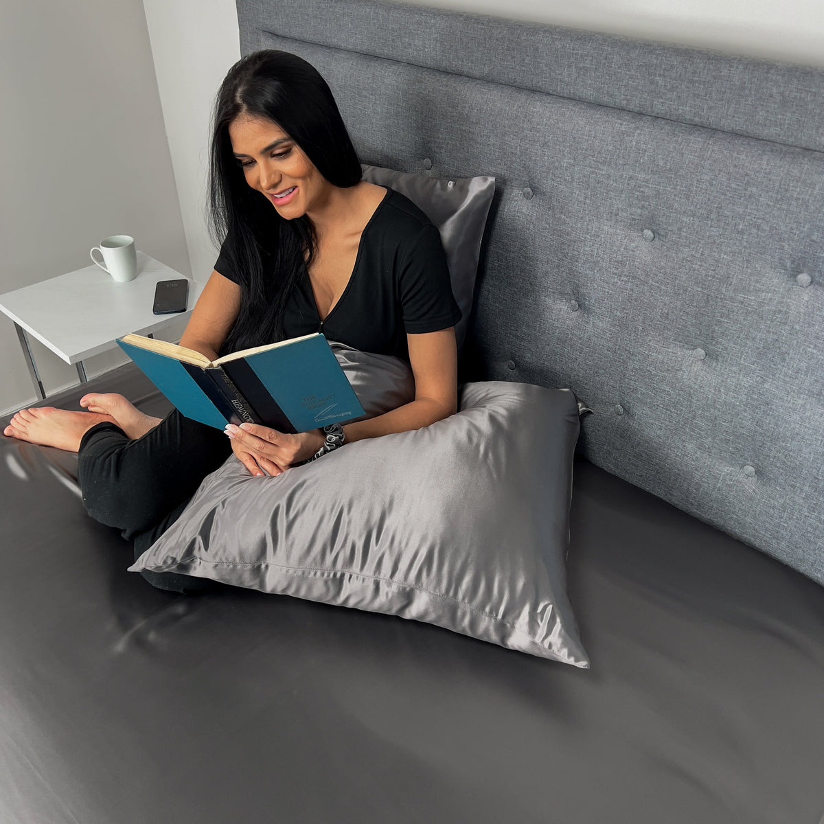 Woman reading and siting on bed with gunmetal silk Euro sham pillow and matching bedding