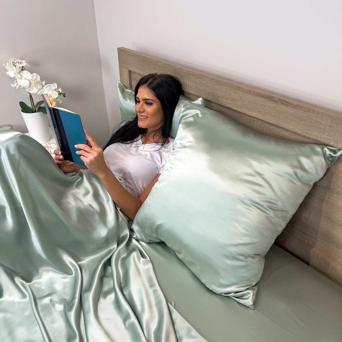 Woman reading and relaxing in bed with Sage silk Euro sham pillow and matching bedding