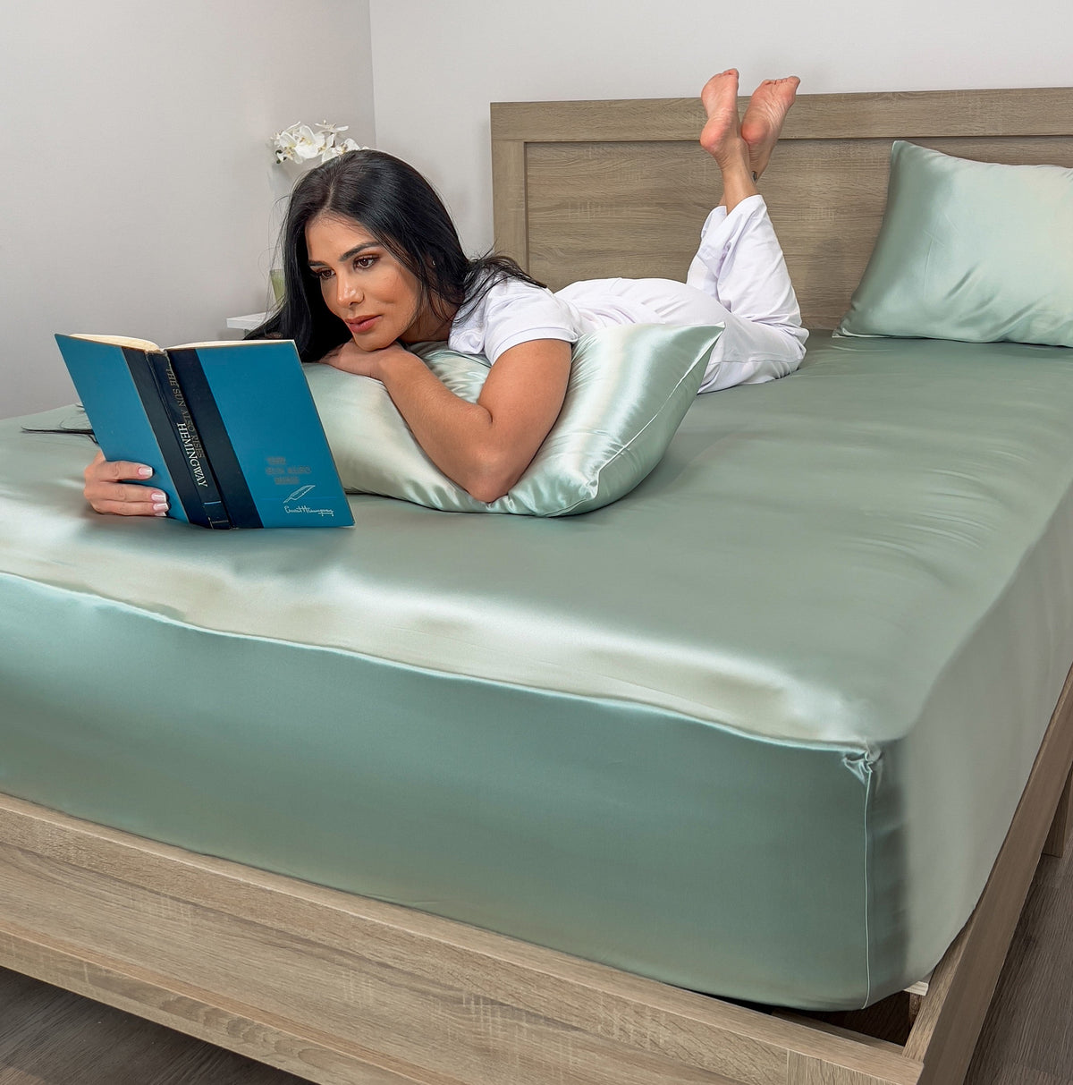 Woman lying on bed with Sage silk fitted sheet and pillow, reading a book and relaxing comfortably