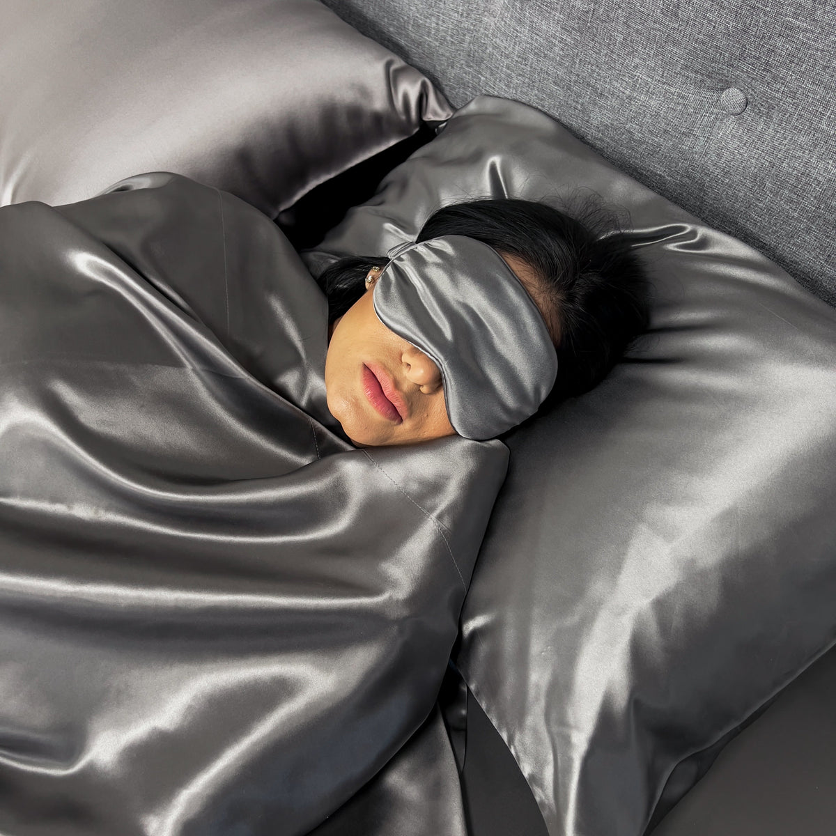 Woman resting on a gunmetal silk pillowcase with a matching sleep mask, lying on the bed.