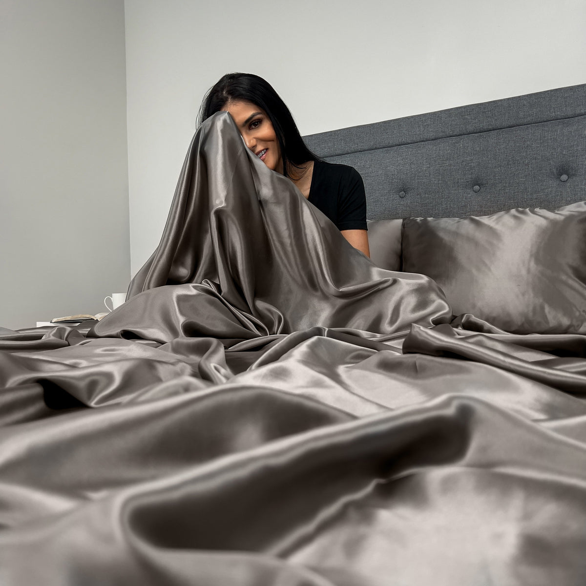Woman sitting on a bed, wrapped in a soft gunmetal silk sheet with a relaxed expression