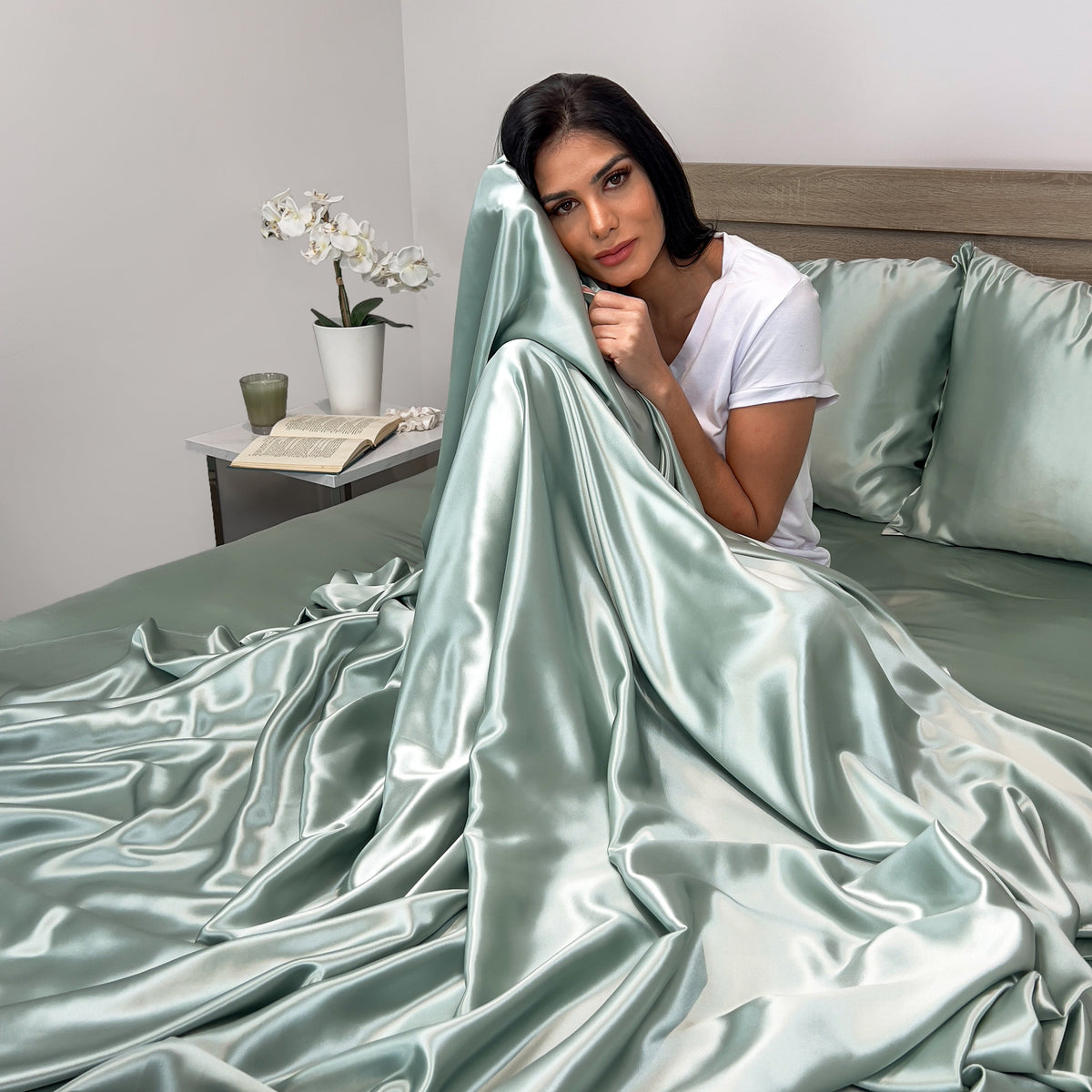 Woman sitting on a bed, wrapped in a soft sage silk sheet with a relaxed expression