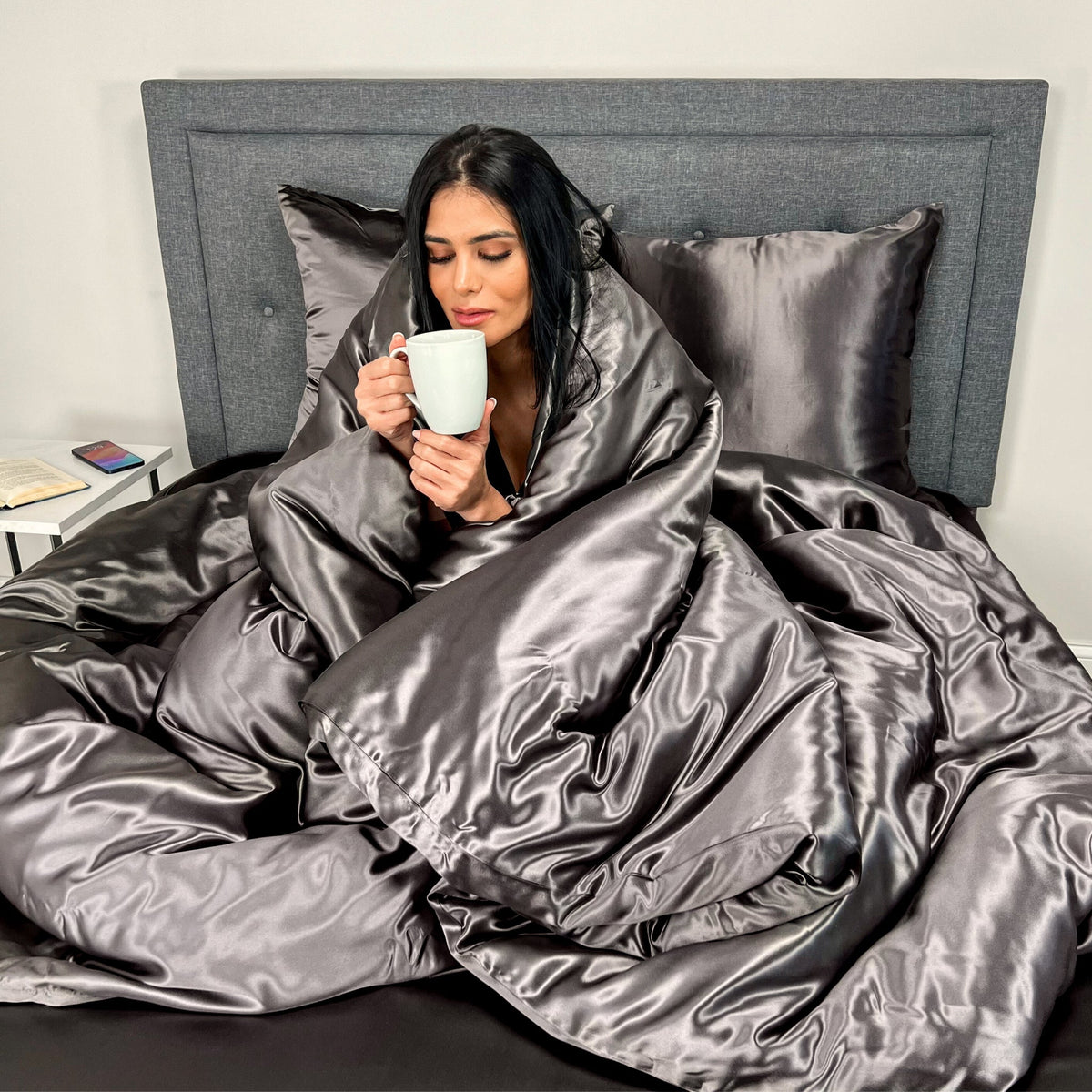 A woman in bed under a silk duvet holding a mug.