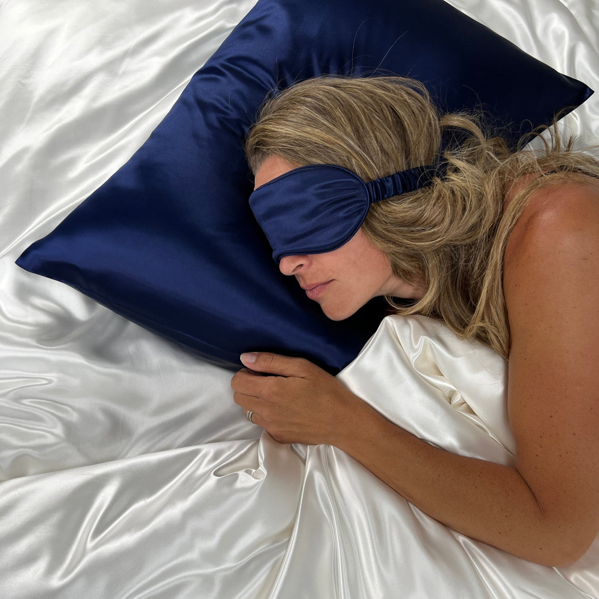 Woman resting on a navy silk pillowcase with a matching sleep mask, sleeping on the bed.