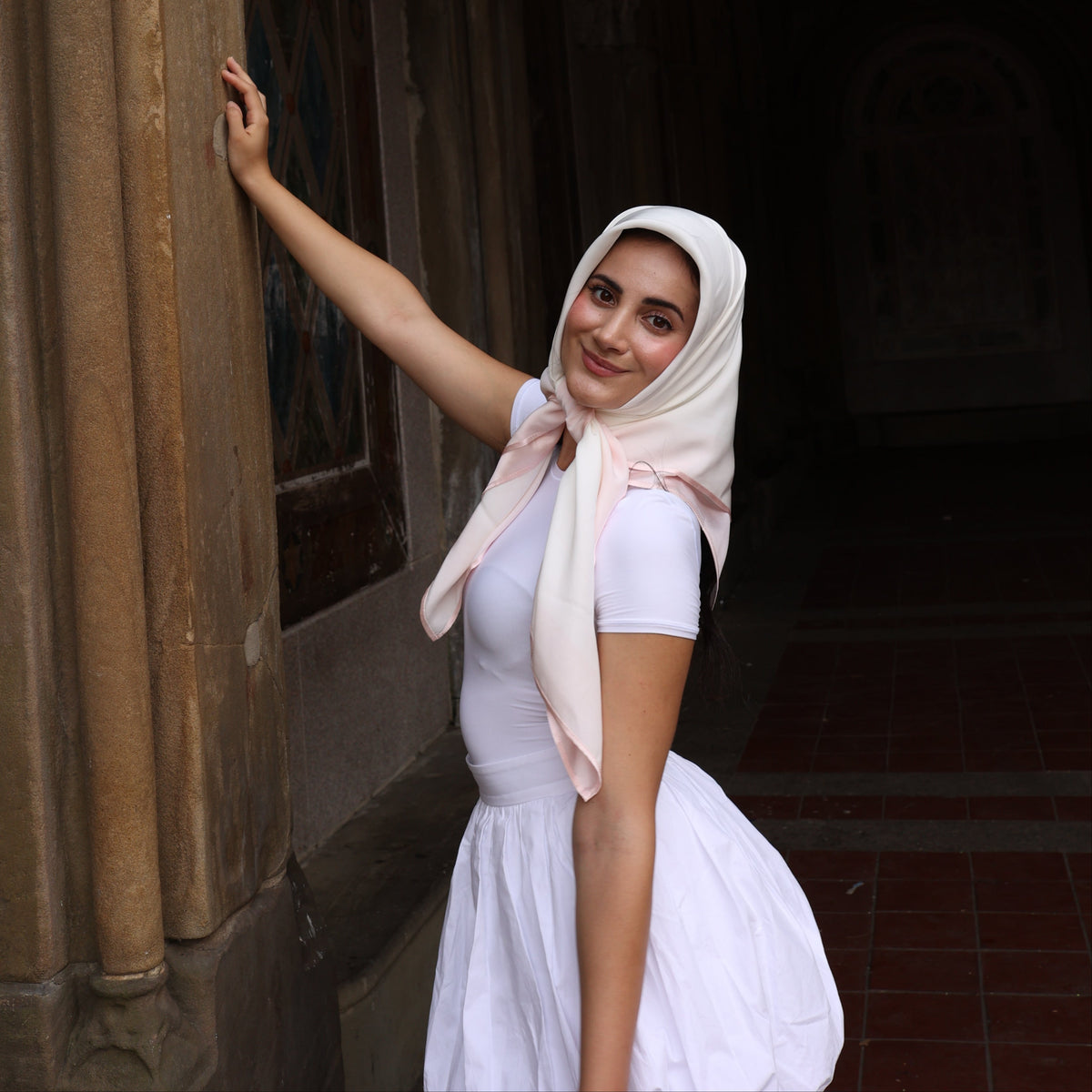 Person wearing a white dress and headscarf standing under a stone archway.