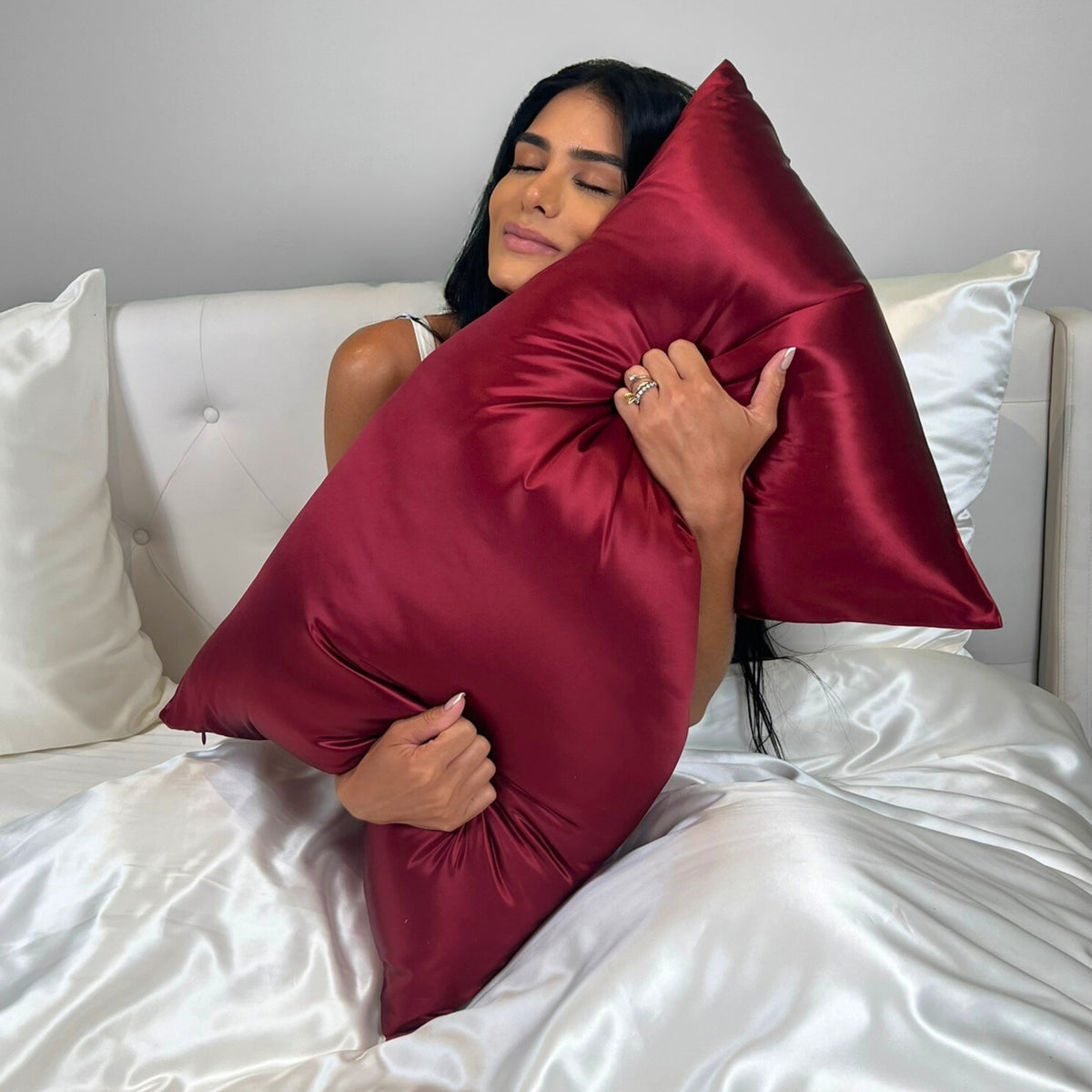 Woman on an ivory silk bed, holding and embracing a cabernet red Mulberry Park Silks pillowcase, highlighting the smooth, lustrous finish of premium Grade 6A silk.