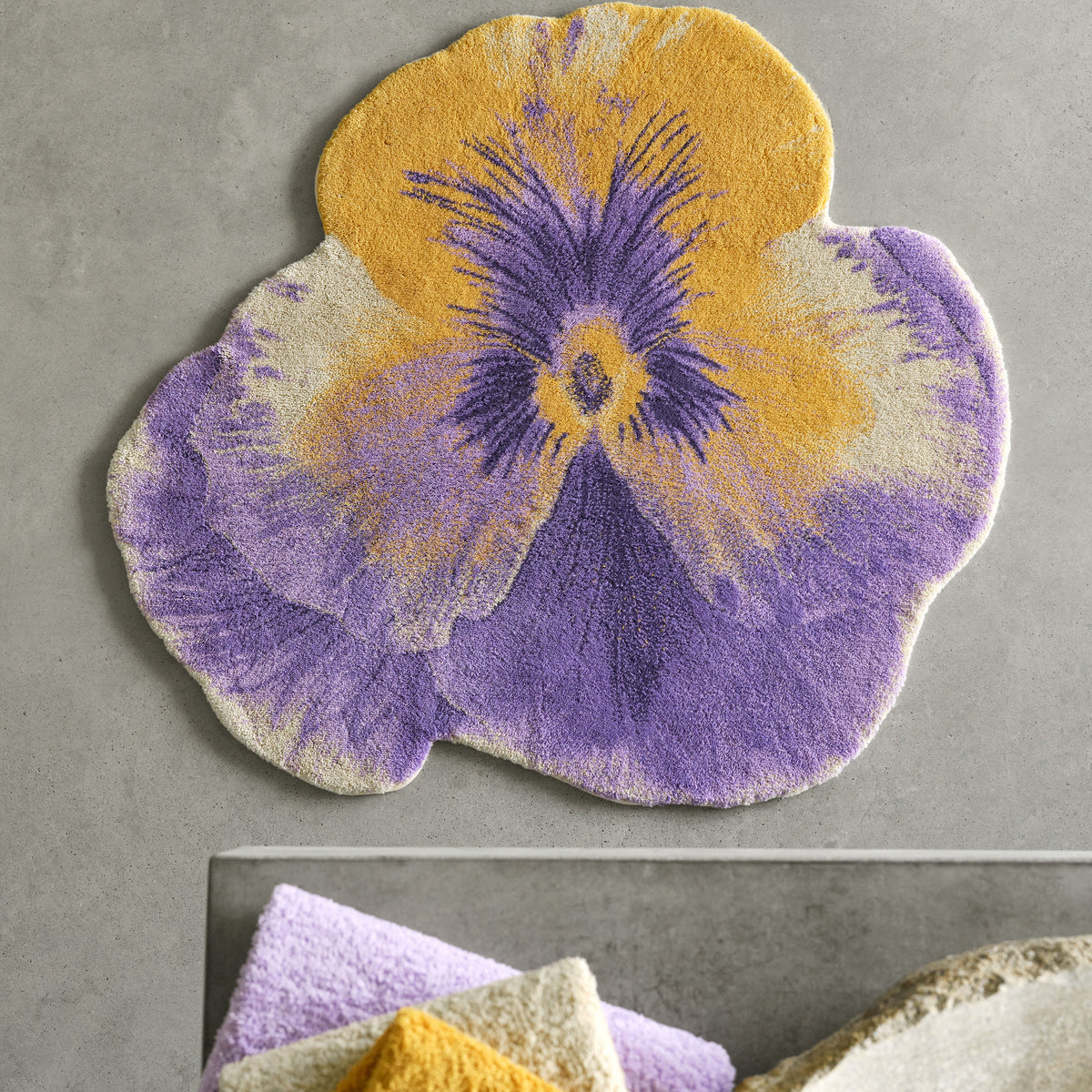 A Pansy Bath Rug by Abyss Habidecor rests on a gray floor beside stacked Egyptian Cotton towels.
