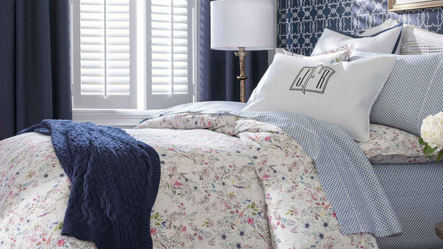 A neatly made bed with floral bedding, a navy knit blanket, monogrammed pillow, and a lamp by a window with shutters.