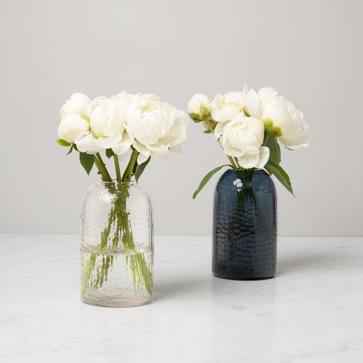 Two glass vases, including a polished glass vase, with white peonies on a white marble surface against a plain background.