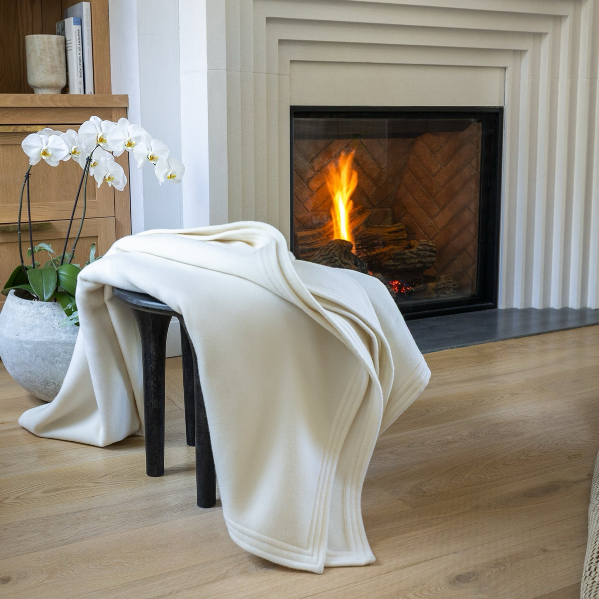 Scandia Home Cortina Cashmere Blanket in Pearl color draped over a dark chair in front of a lit fireplace with a potted orchid nearby