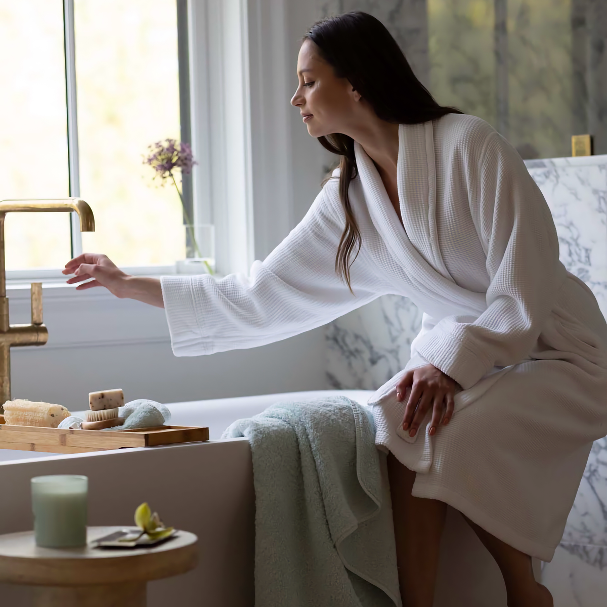 Woman in a White Scandia Home Grotto Robe sitting by a bathtub with a spa-like setting.