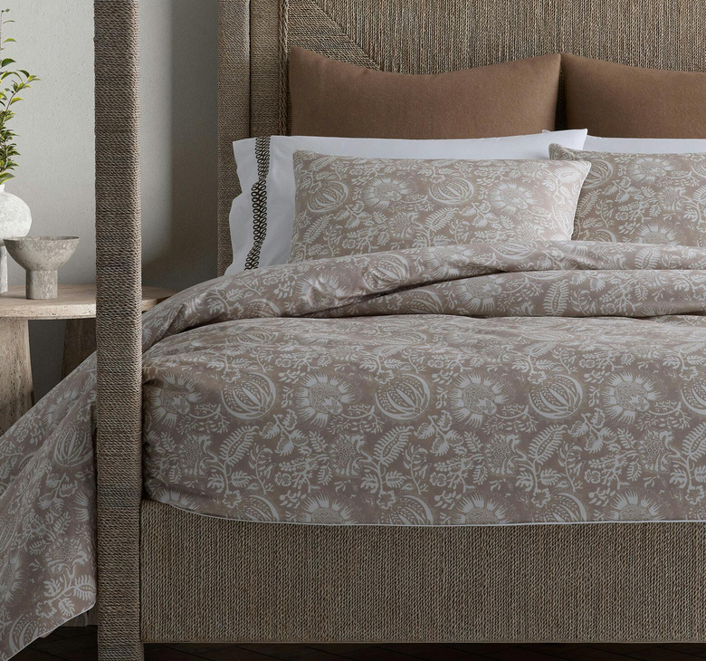 A bed with a beige floral comforter, matching pillows, and a textured headboard next to a wooden nightstand.