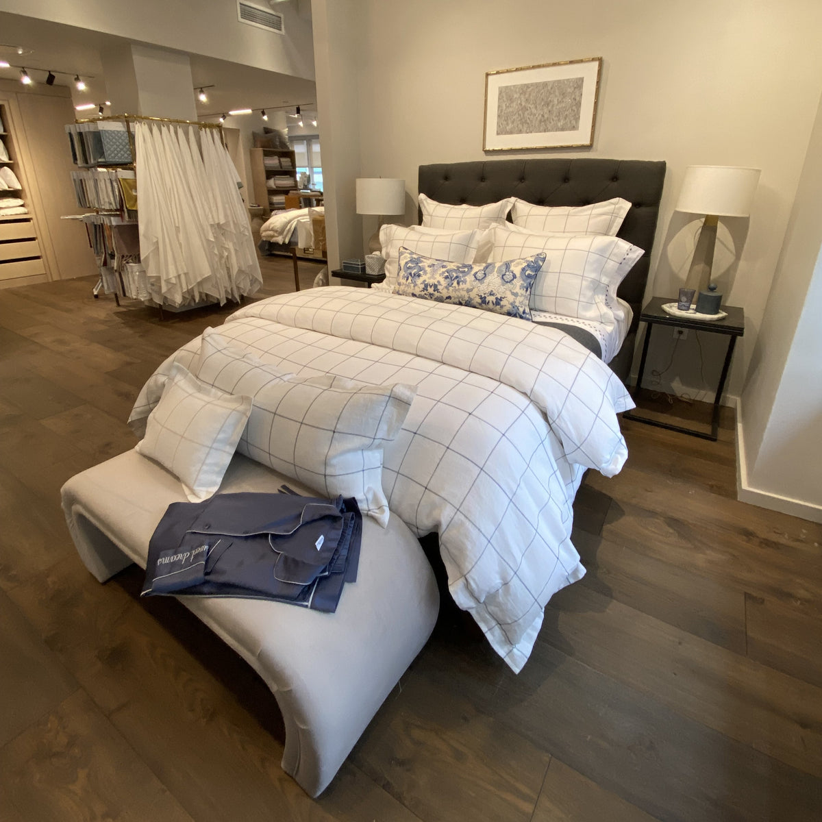 A neatly made bed with Matouk Sebastian Bedding and square-patterned embroidery is displayed with lamps, benches, and linens.