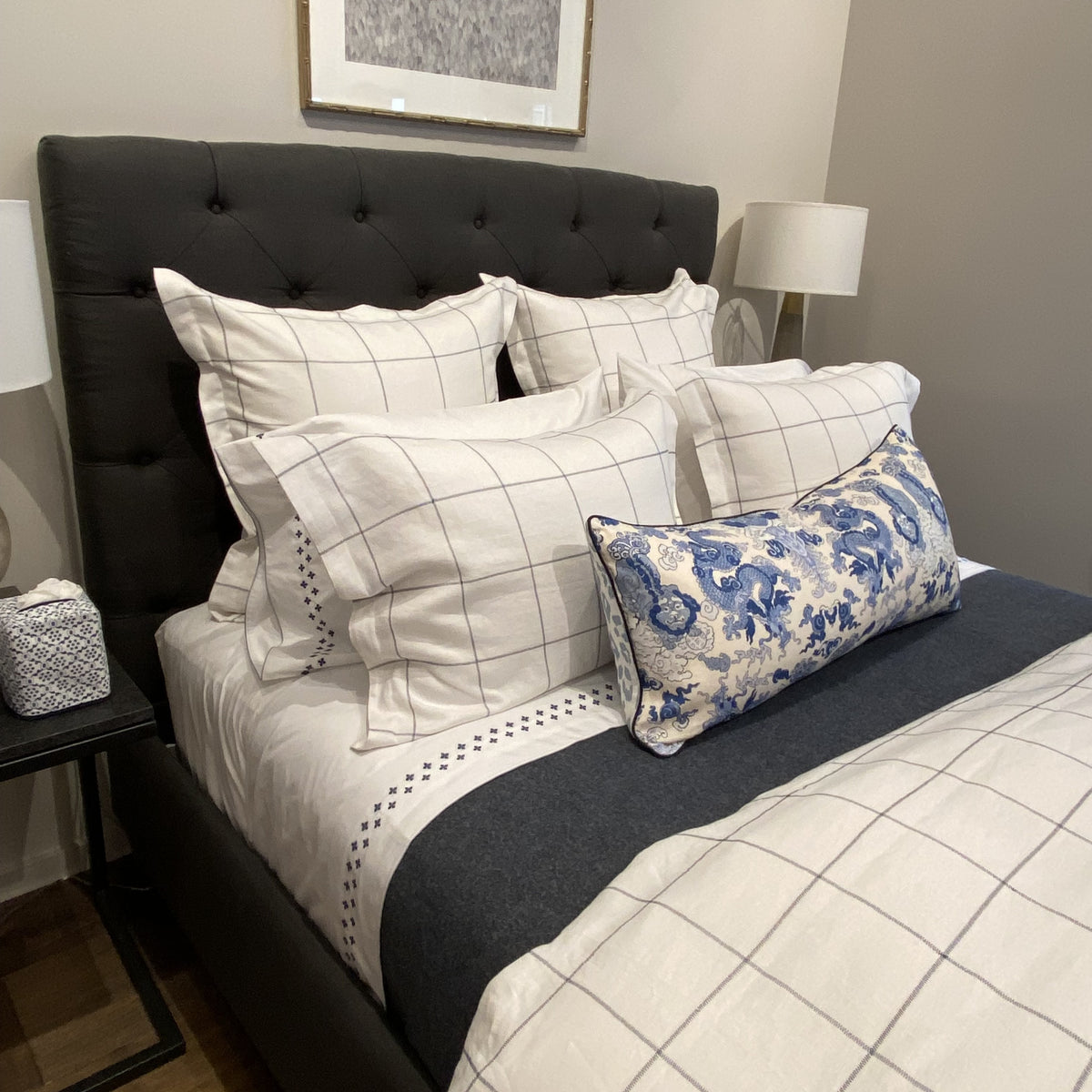 A neatly made bed with Matouk Sebastian Bedding, blue floral accent pillow, and a dark upholstered headboard.