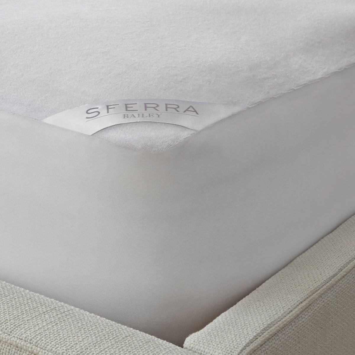 Close-up of a SFERRA BAILEY cotton terry mattress protector fitted over a mattress on a beige bed frame.