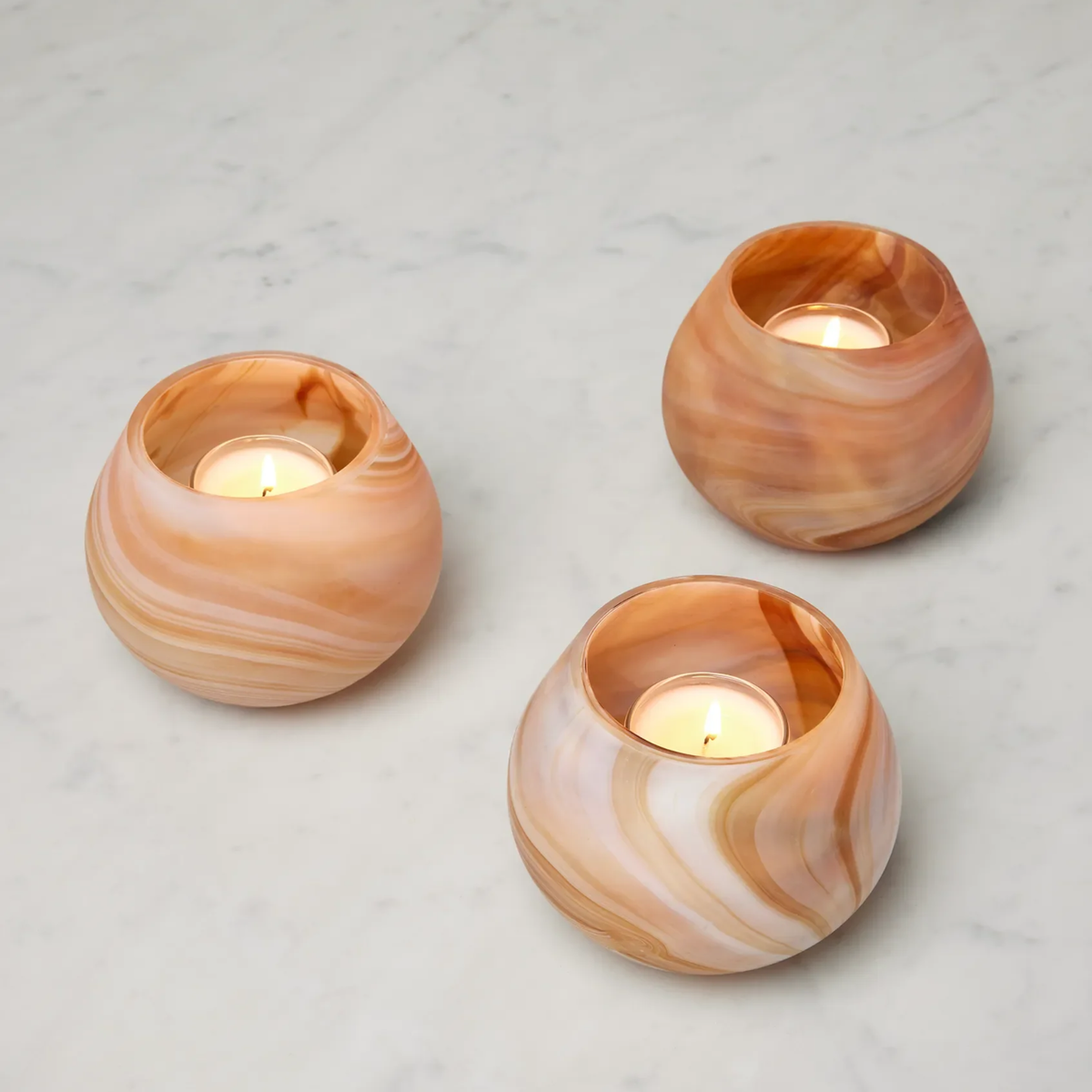 Three round, Almond Sferra Giove Marbled Votive Holders with lit tealights add unique home decor charm to a light marble surface.