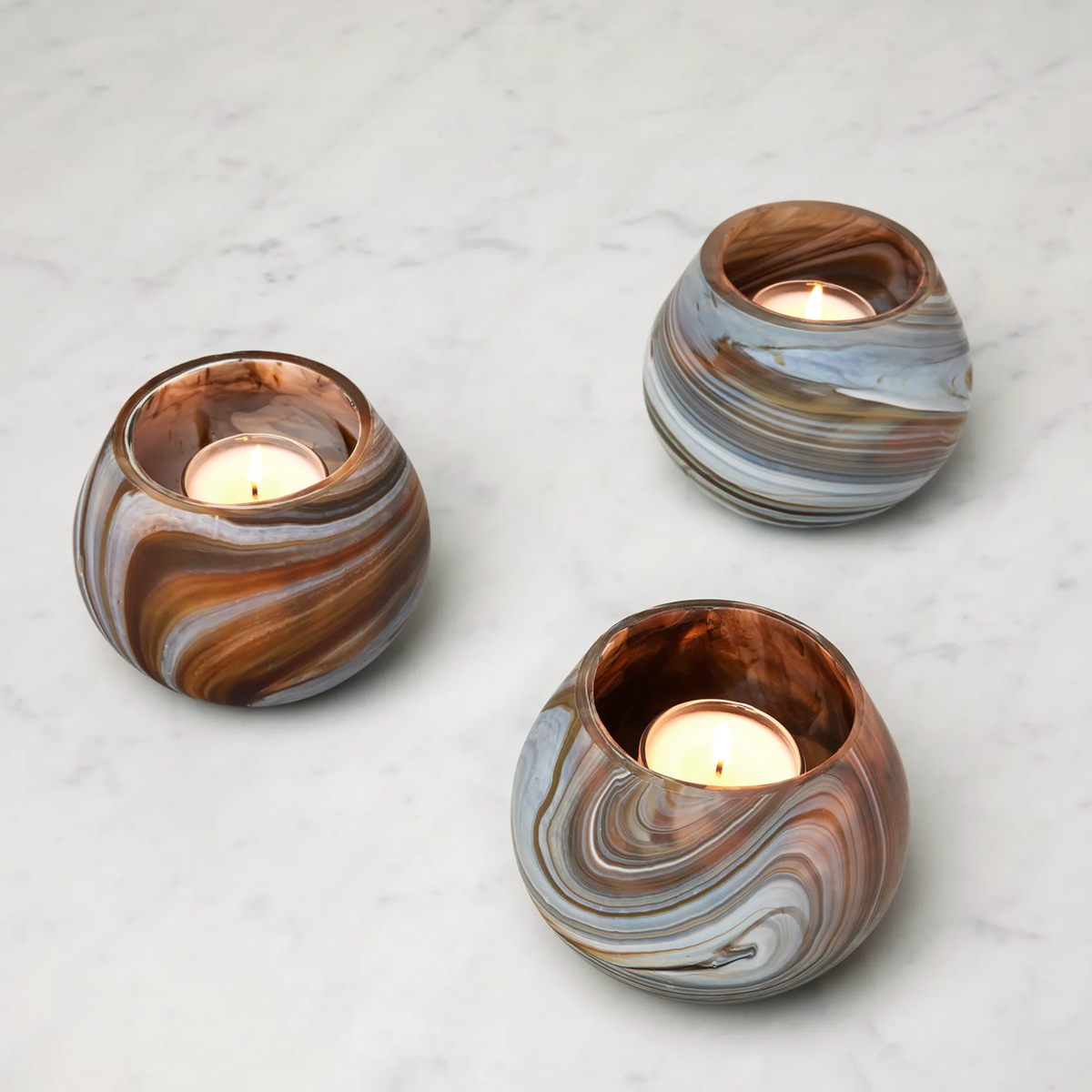 Three round, Bark Sferra Giove Marbled Votive Holders with lit candles create unique home decor on a white marble surface.