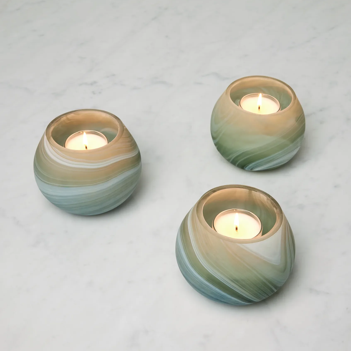 Three lit Clover Sferra Giove Marbled Votive Holders glow on a light marble surface.