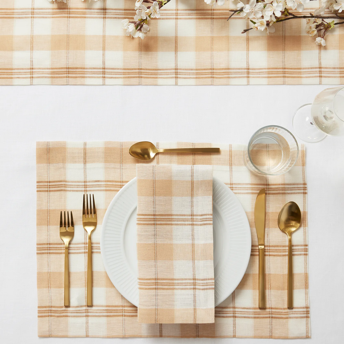 A table setting with gold utensils, a white plate, Sferra Masseria Table Linens in Almond and Oyster colors, glass of water, and flowers