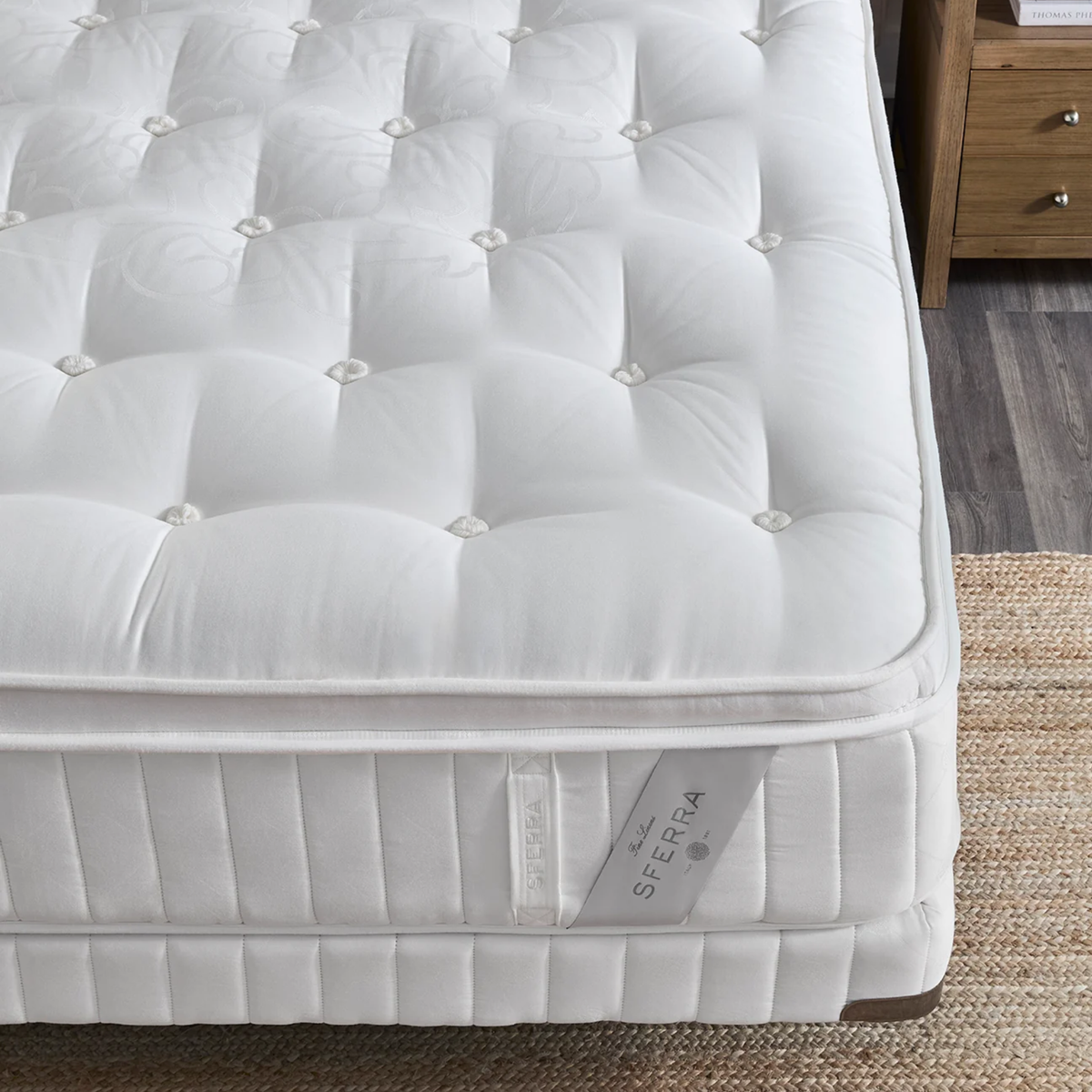 White, quilted luxury Sferra Nuvole CV Pillowtop Mattress with button tufting on wood floor near brown nightstand and a woven beige rug.
