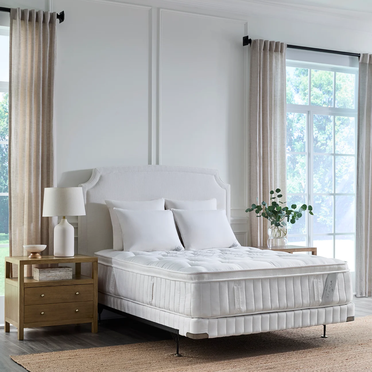 A neatly made bed with Sferra Nuvole CV Pillowtop Mattress, wooden nightstand, lamp, and greenery in a bright bedroom.