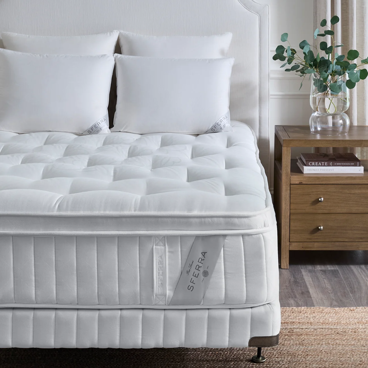 A neatly made bed with white pillows and an Sferra Nuvole CV Pillowtop Mattress, beside a nightstand with a vase and books.