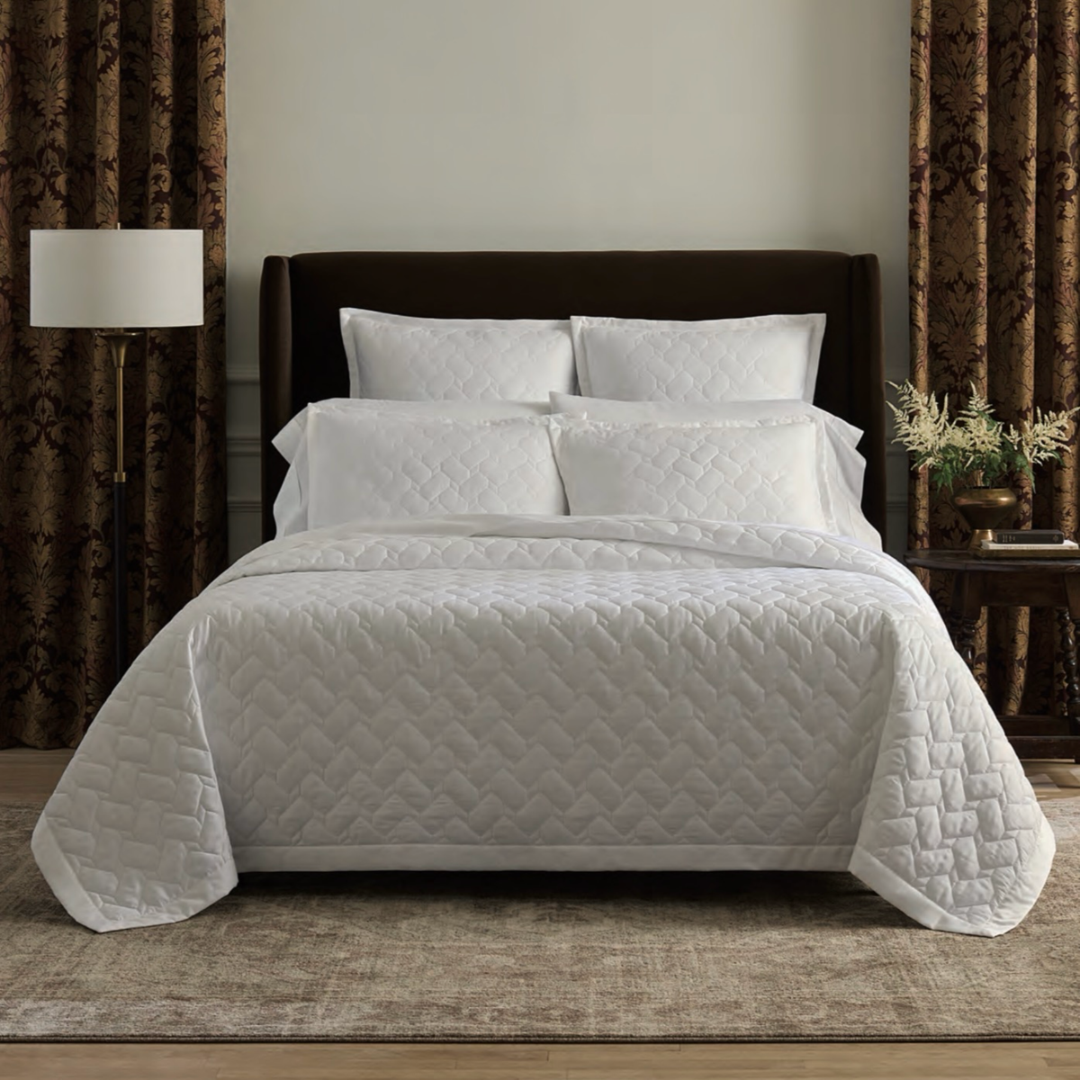 A neatly made bed with White Sferra Palatino Quilt and Shams, against a dark headboard, flanked by brown curtains.