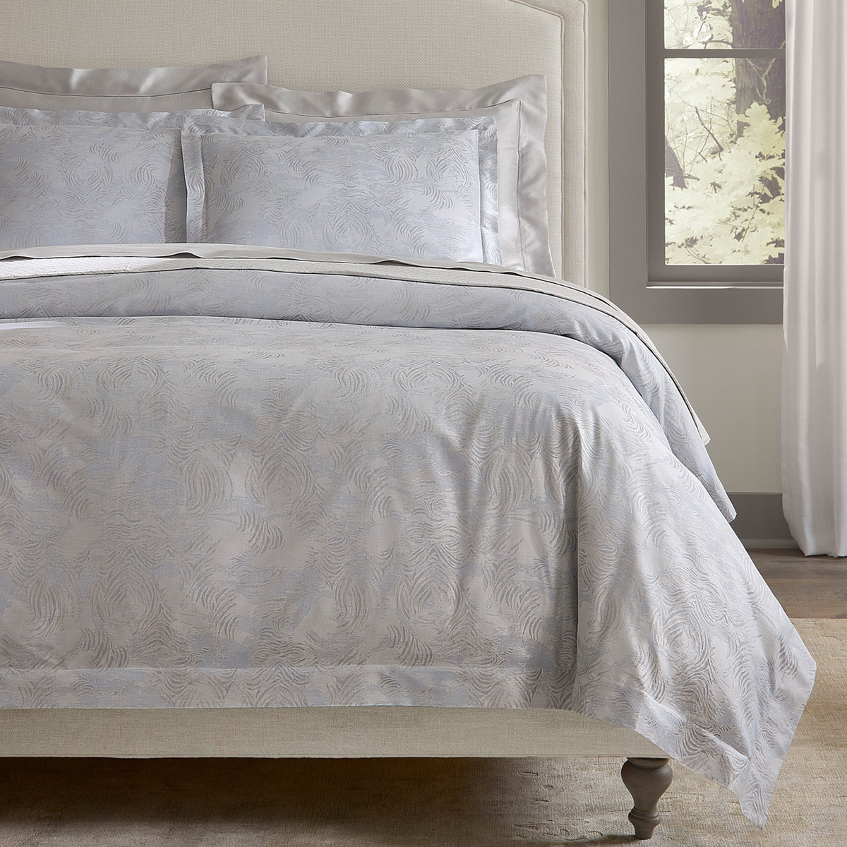 A neatly made bed features a Sferra Riflesso duvet cover, crafted from long-staple cotton, by a window with white curtains.