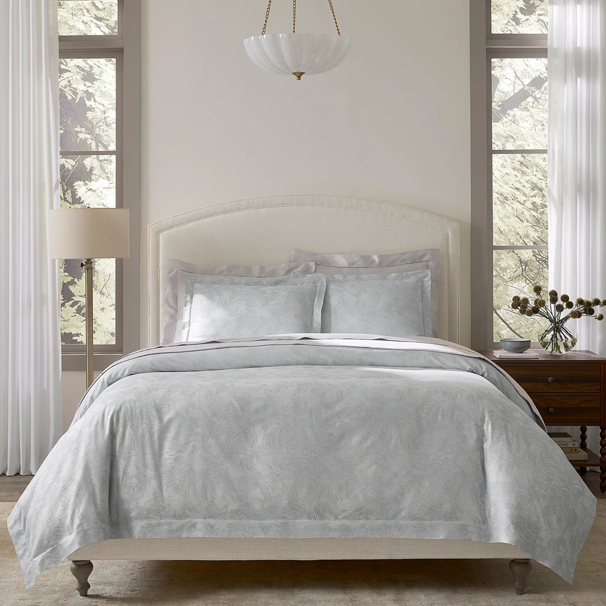 A neatly made bed with a Sferra Riflesso duvet cover, two pillows, and a lamp in a bright room with large windows.